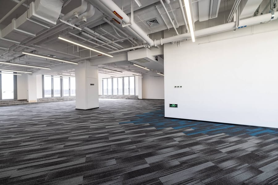 Large open-plan commercial floor laid with grey plank-style carpet tile and a blue accent run, over a raised subfloor