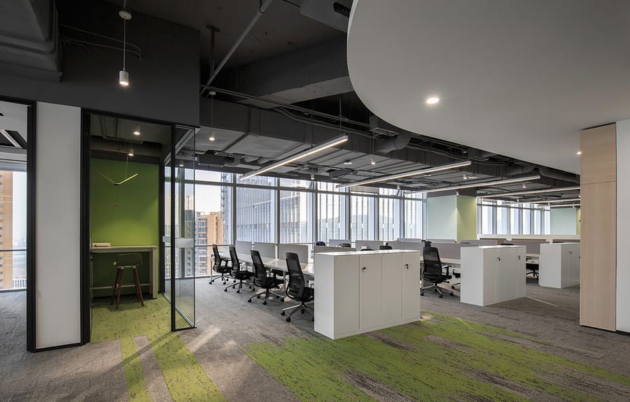 Open-plan office with workstations and grey carpet tile broken by a green accent path, over an exposed-services ceiling