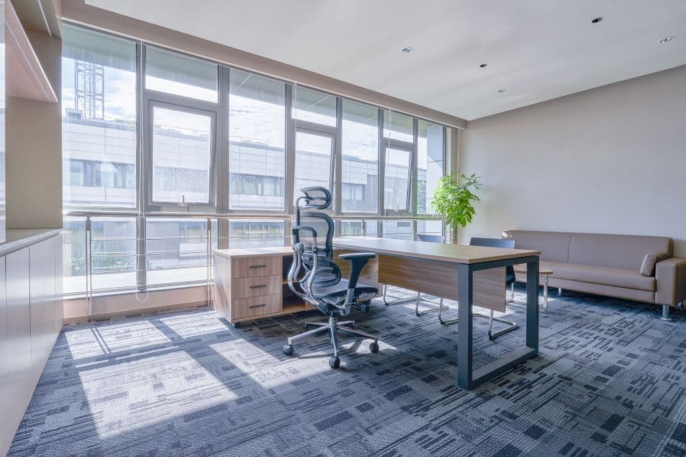 Open-plan office with grey patterned modular carpet tile, glass curtain wall and hard ceiling — the reflective surfaces that make floor absorption matter